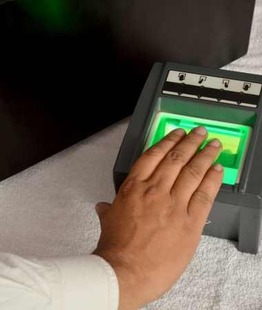 A person is placing their hand on a live scan fingerprinting machine for biometric identification. The hand is illuminated by a green light emitted from the rectangular device, which features several icons along the top edge and rests on a white cloth surface.