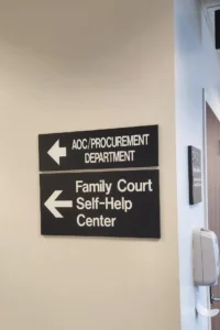 A hallway with two black signs mounted on a white wall. The upper sign, with a left-pointing arrow, reads "AOC/Procurement Department." The lower sign, also with a left-pointing arrow, reads "Family Court Self-Help Center," offering assistance for Adult Name Change and Live Scan Fingerprinting services. A hand sanitizer dispenser is mounted on the wall to the right.